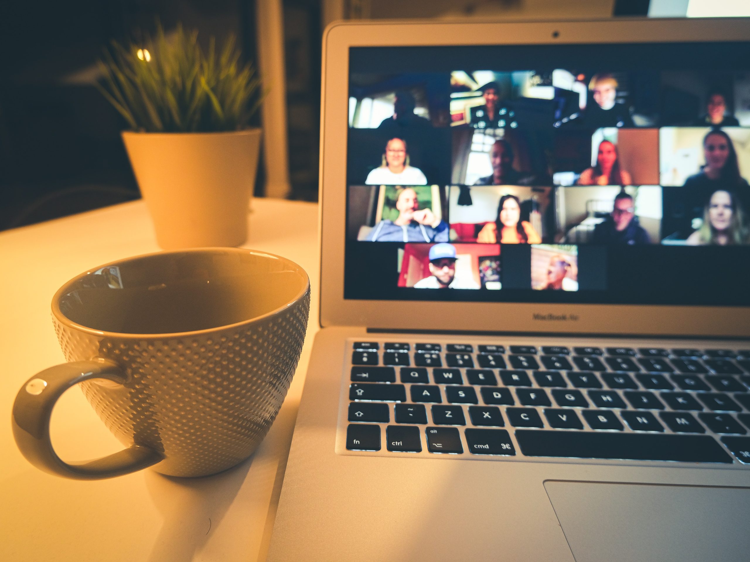 Photograph of a laptop screen showing multiple people on a video call, next to the laptop is a mug