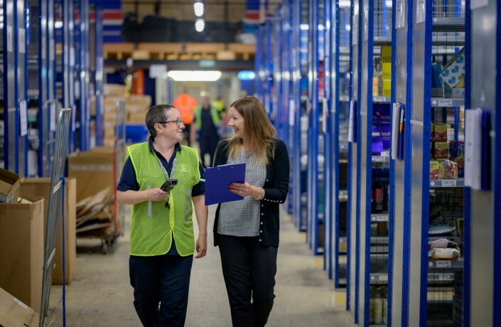 Two women walking through a warehouse and talking. One is holding a clipboard.