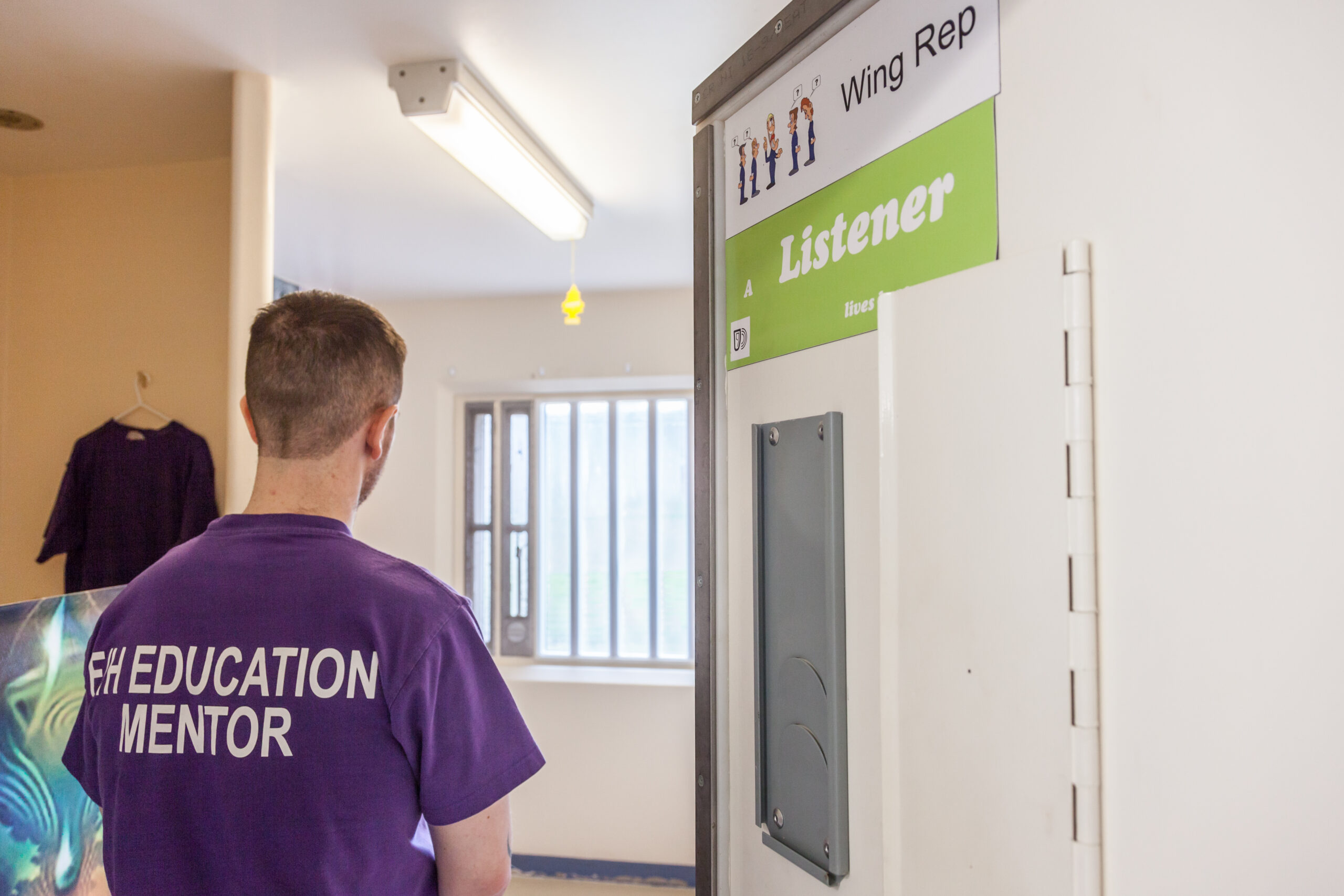 Learner wearing a F/H Education Mentor t-shirt walks into his cell, which has signs saying Wing Rep and Listener on the door